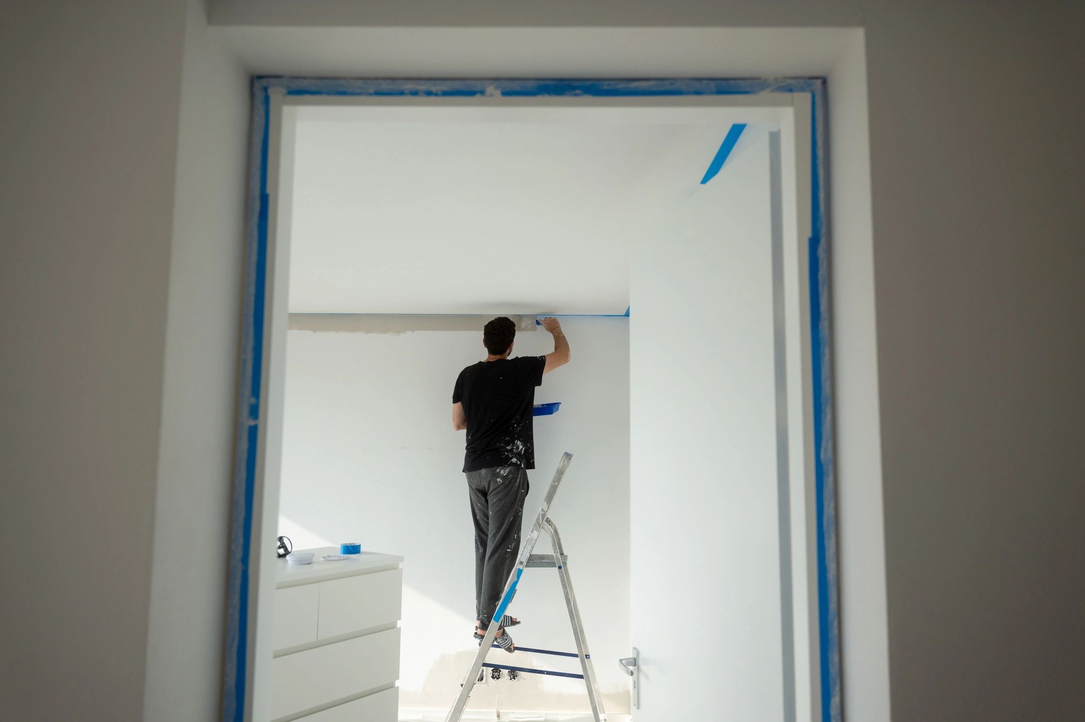 Person painting a white wall standing on a ladder inside a room.