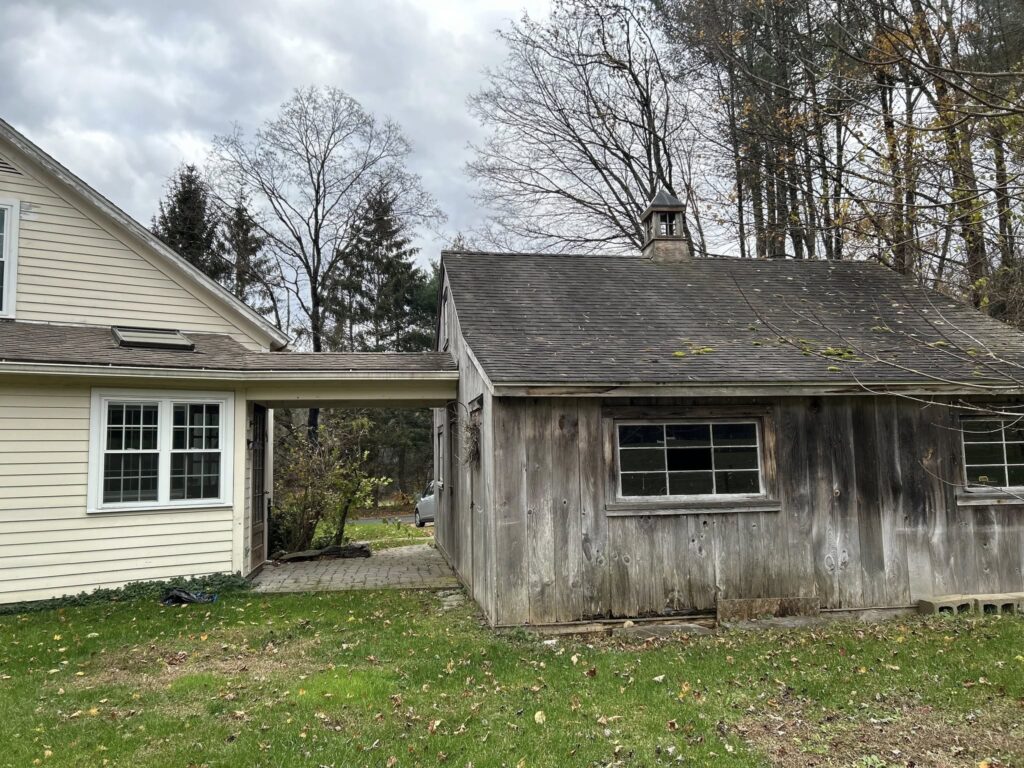 A rustic wooden shed next to a white house under a cloudy sky.